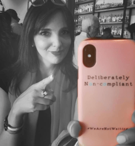 Black and white photo of me pointing at the camera. I'm holding up my phone case which is highlighted in pink and has the words 'Deliberately non-compliant' printed in black.