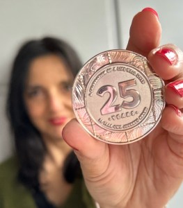 Photo of me holding a silver coin. The  coin has the number 25  stamped on it and I’m holding it out in front of me. I’m blurred in the background.